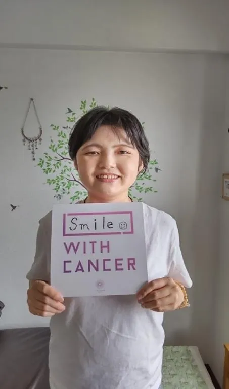 「Smile WITH CANCER」と書いたボードを掲げて微笑む園田彩夏さん