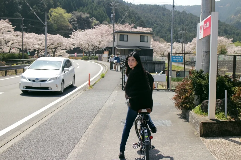 桜並木の道で自転車にまたがり振り返る友美さん